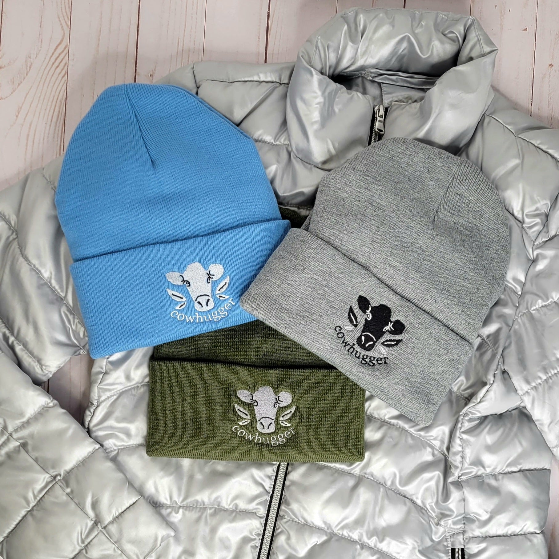 Three embroidered Cowhugger cuffed beanies lying on a winter puffer jacket.  Light blue beanie with white logo, gray beanie with black and white logo, and military green beanie with silver logo.